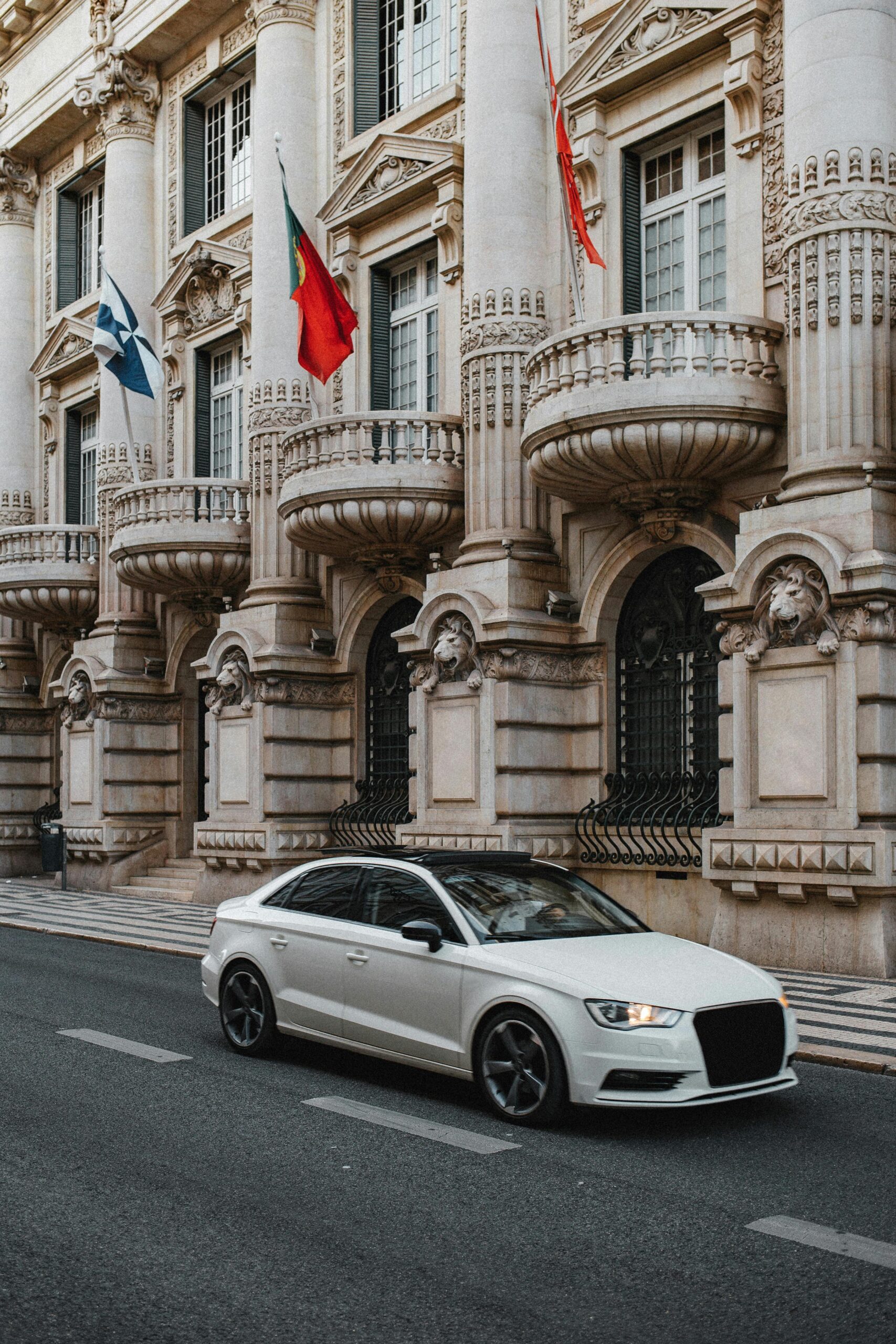 A stylish white sedan driving past a historic European architectural facade on a city street.